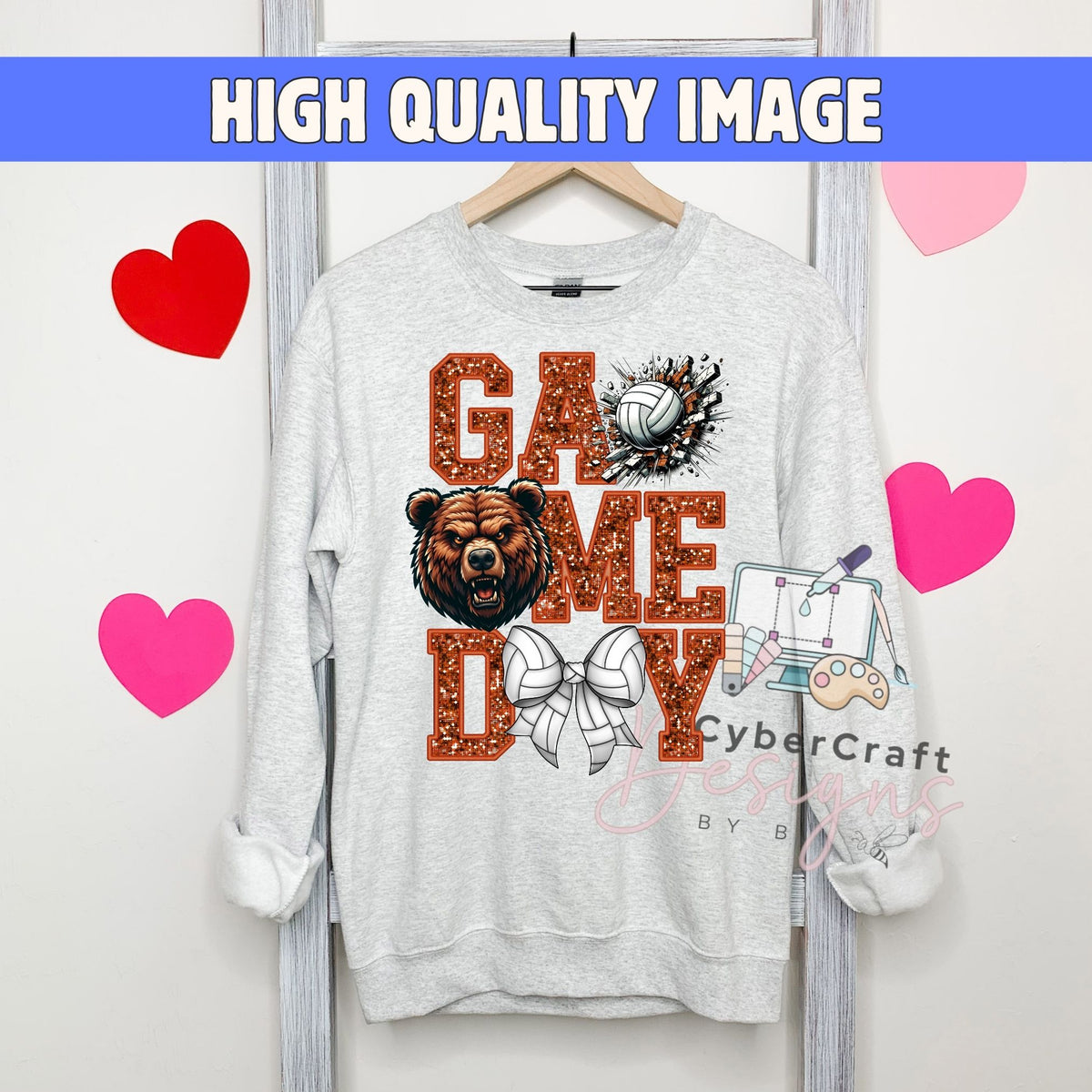A light gray crewneck sweatshirt hanging on a wooden hanger, featuring a 'GAME DAY' graphic with a brown bear, volleyball, and sports illustrations. The image has a blue banner at the top reading 'HIGH QUALITY IMAGE' and pink heart graphics on the sides.
