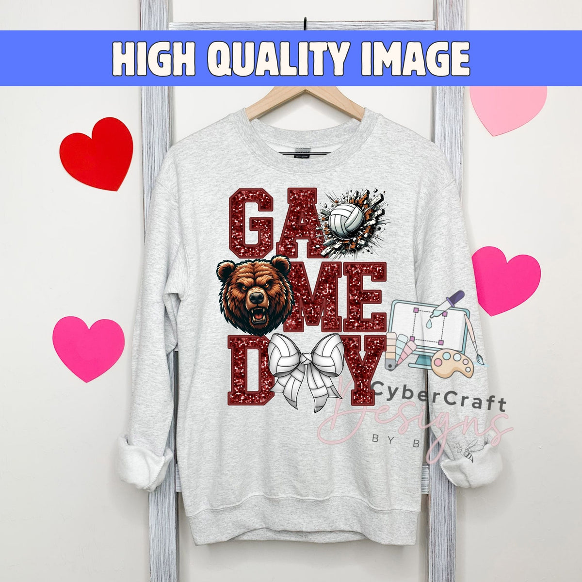A light gray crewneck sweatshirt hanging on a wooden hanger, featuring a 'Game Day' graphic with a bear, volleyball, and bow tie. The image has a blue banner at the top reading 'HIGH QUALITY IMAGE' and pink heart graphics on the sides.